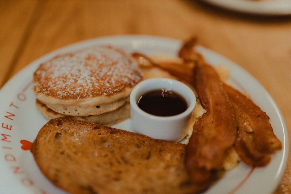 A close-up of a breakfast plate featuring pancakes, bacon, toast, and syrup.