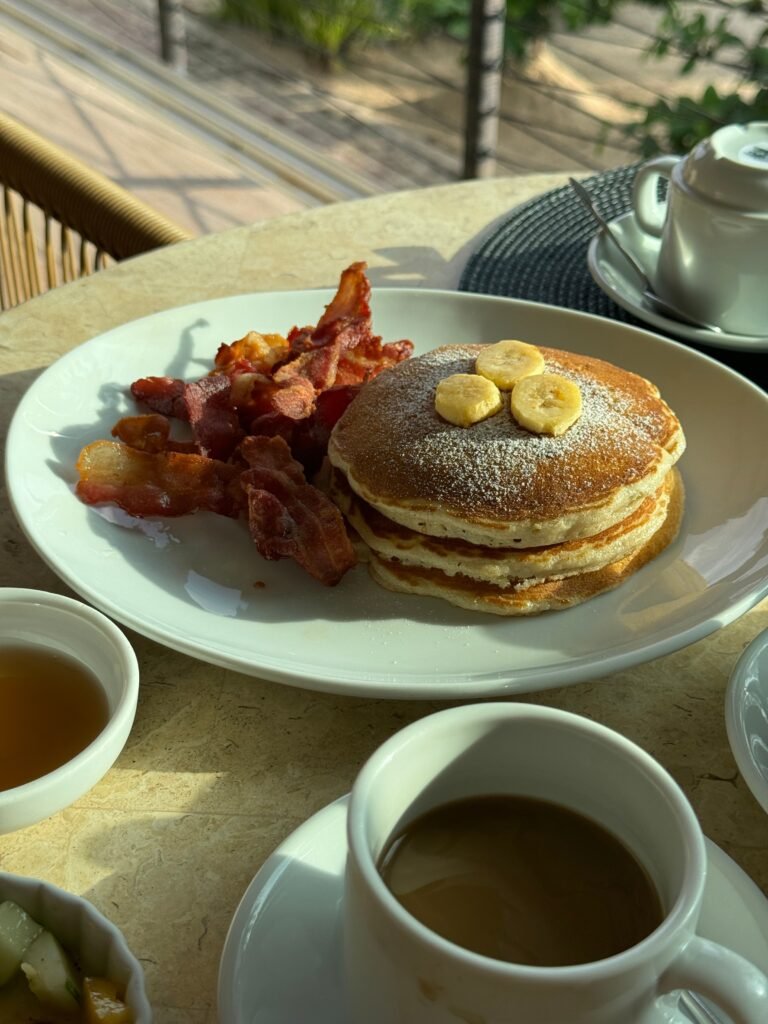A tasty breakfast spread with pancakes, banana slices, bacon, and coffee on a sunny morning.