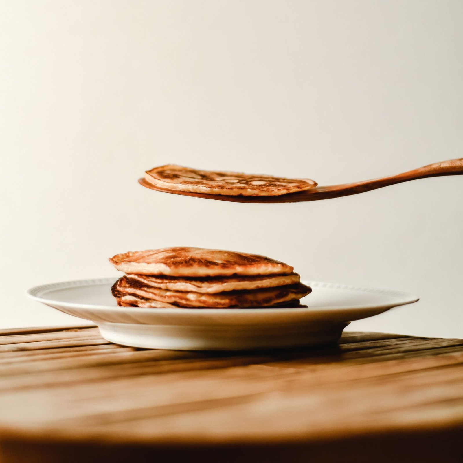 A stack of golden pancakes on a plate with one being lifted by a spatula, indoor setting.